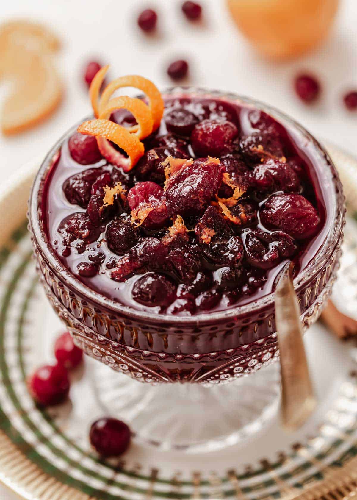 cranberry sauce in a glass compote sitting on fancy plate with cranberries and oranges around.