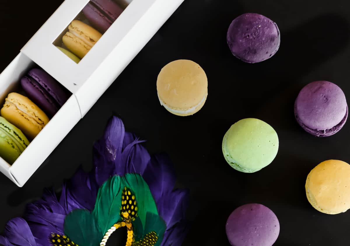 black backdrop with purple, gold, and green macarons with favor box and mardi gras mask, overhead view.