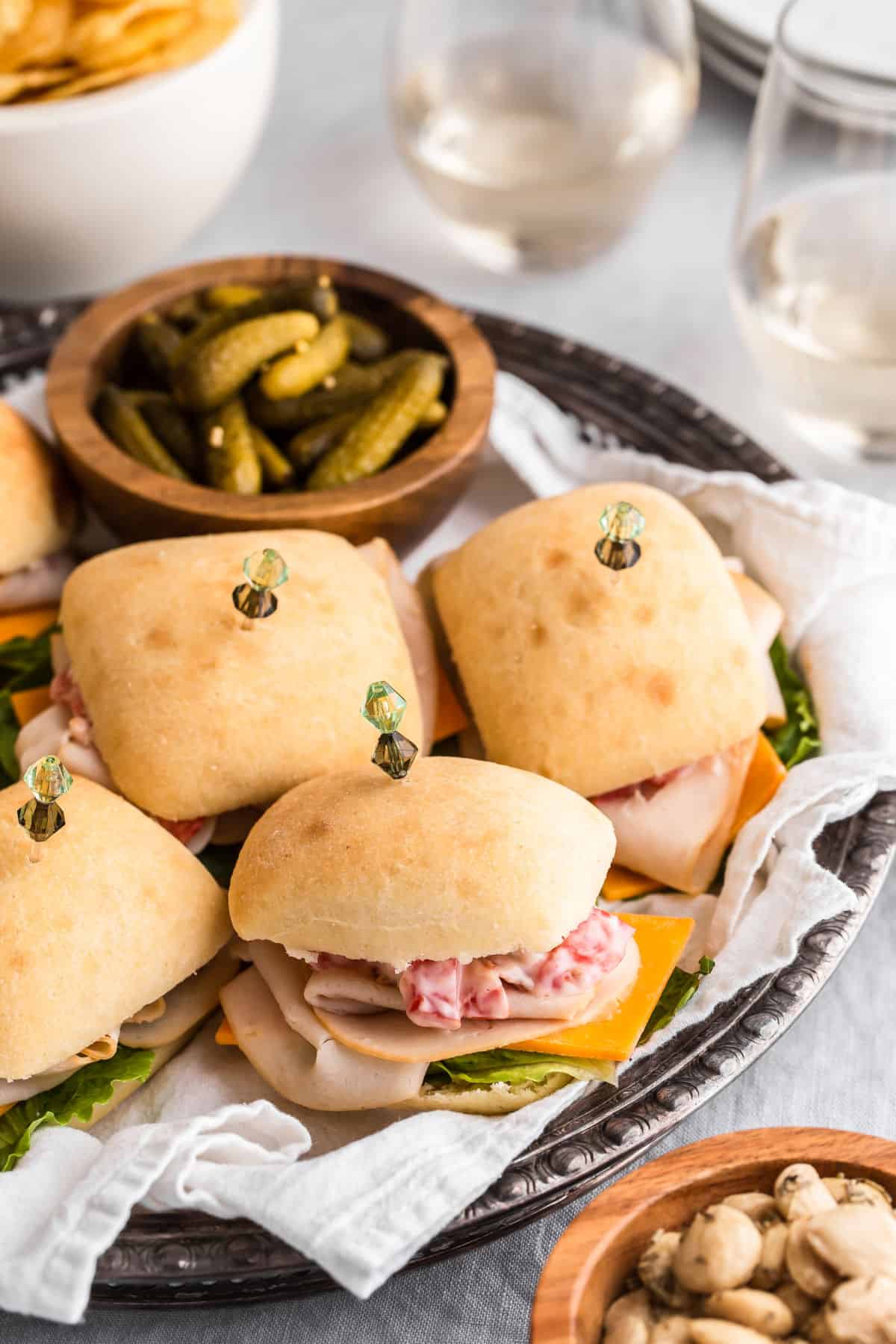 mini party sandwiches on white cloth lines silver tray, served with pickles and almonds.