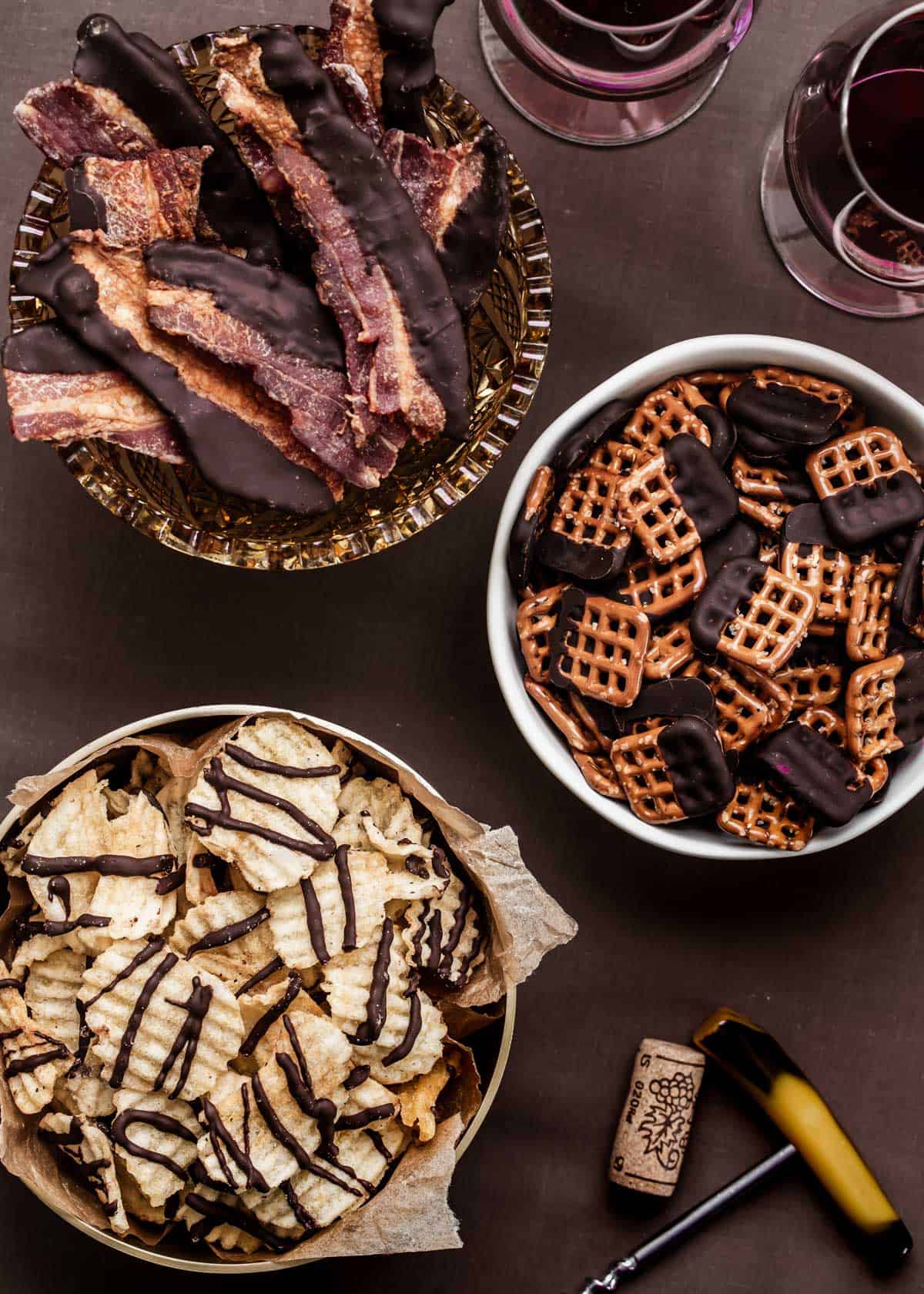 chocolate dipped bacon, pretzels, and potato chips in bowls on brown table.