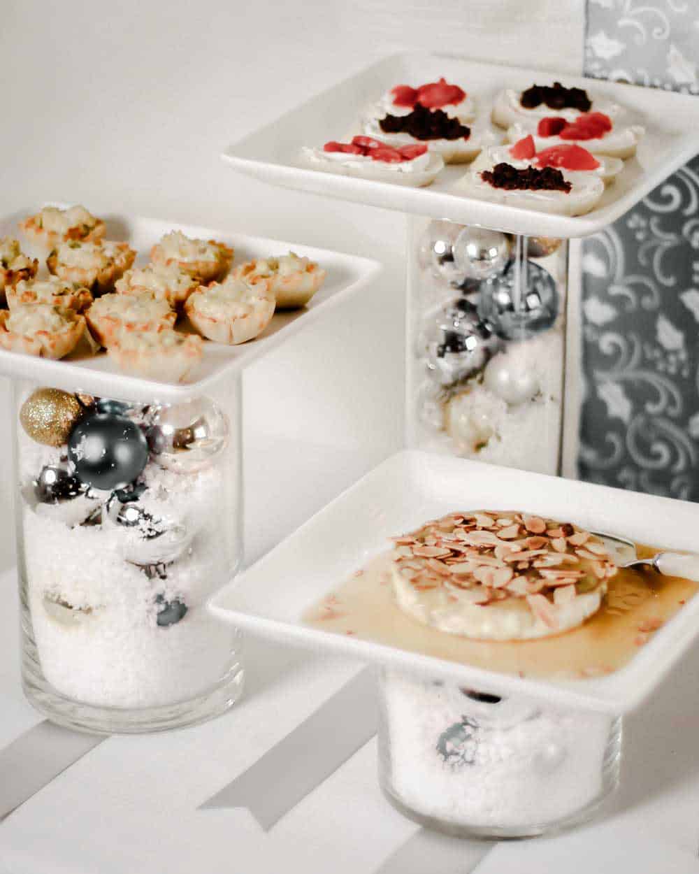 three clear vases filled with Christmas ornaments, with white plates of food on top.