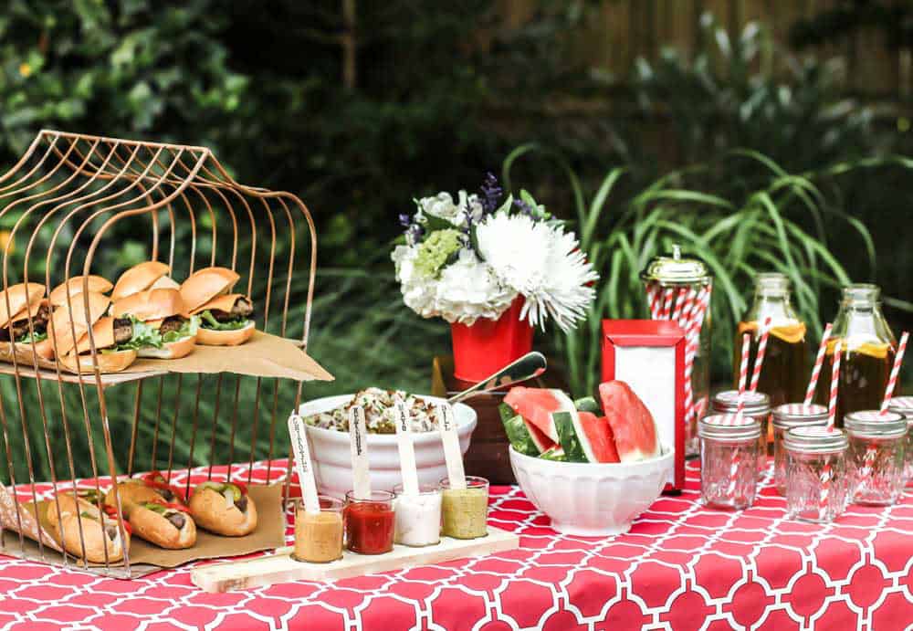 summer cookout buffet table set up outside on red tablecloth.