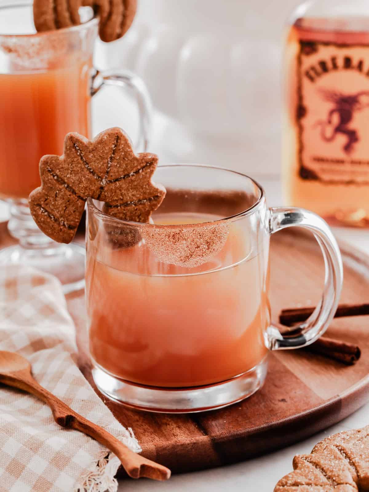 hot cider garnished with cookie on rim of glass mug, with Fireball bottle in background.