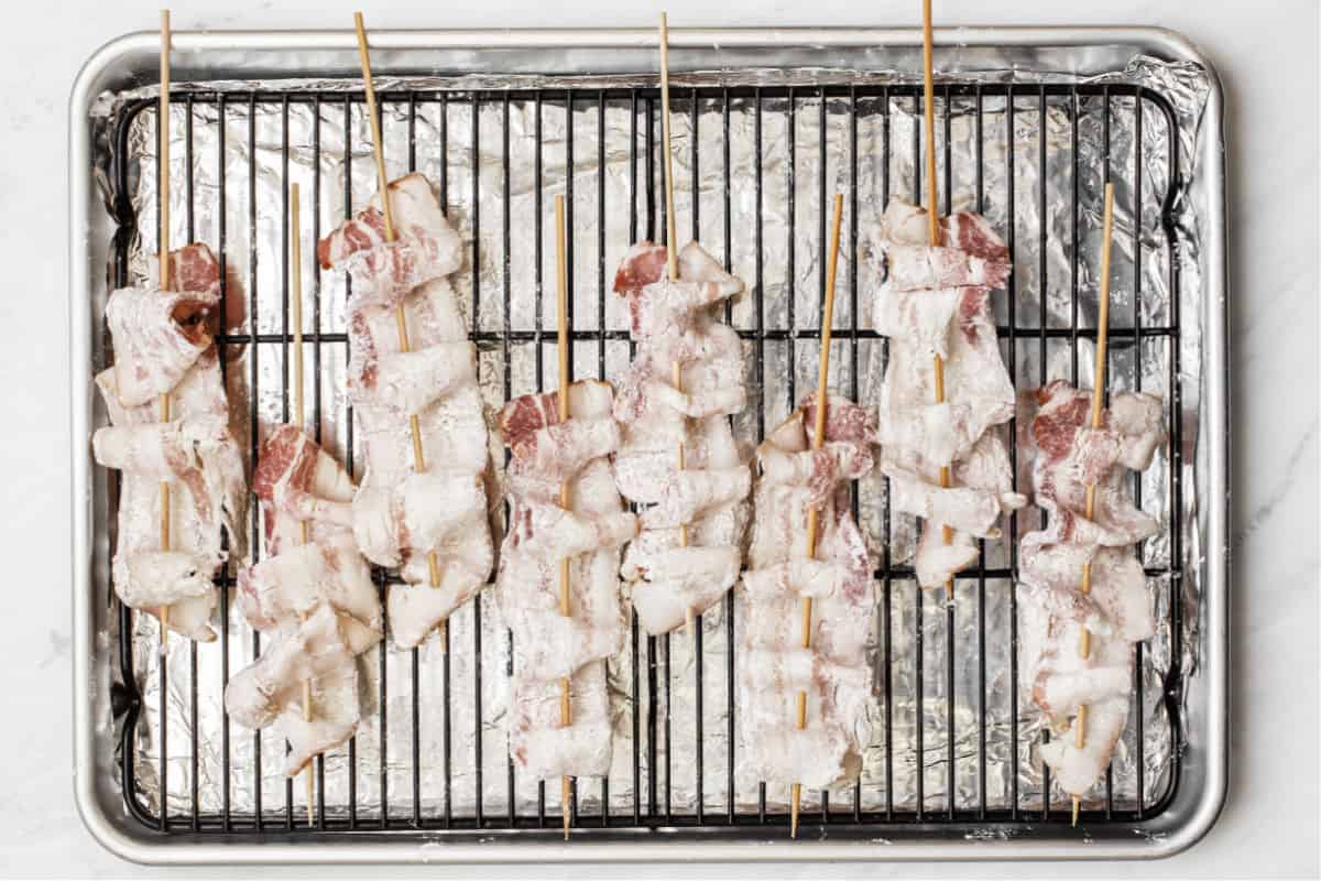 raw bacon on wood skewers laying on baking rack on sheet pan, overhead.