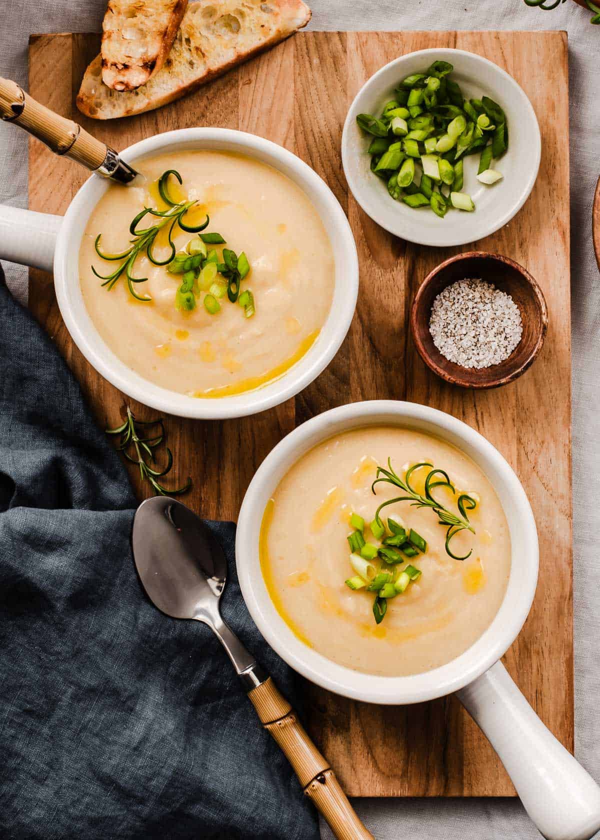 two bowls of smooth white soup, sitting on wood board with crostini bread and bowl of diced green onions.