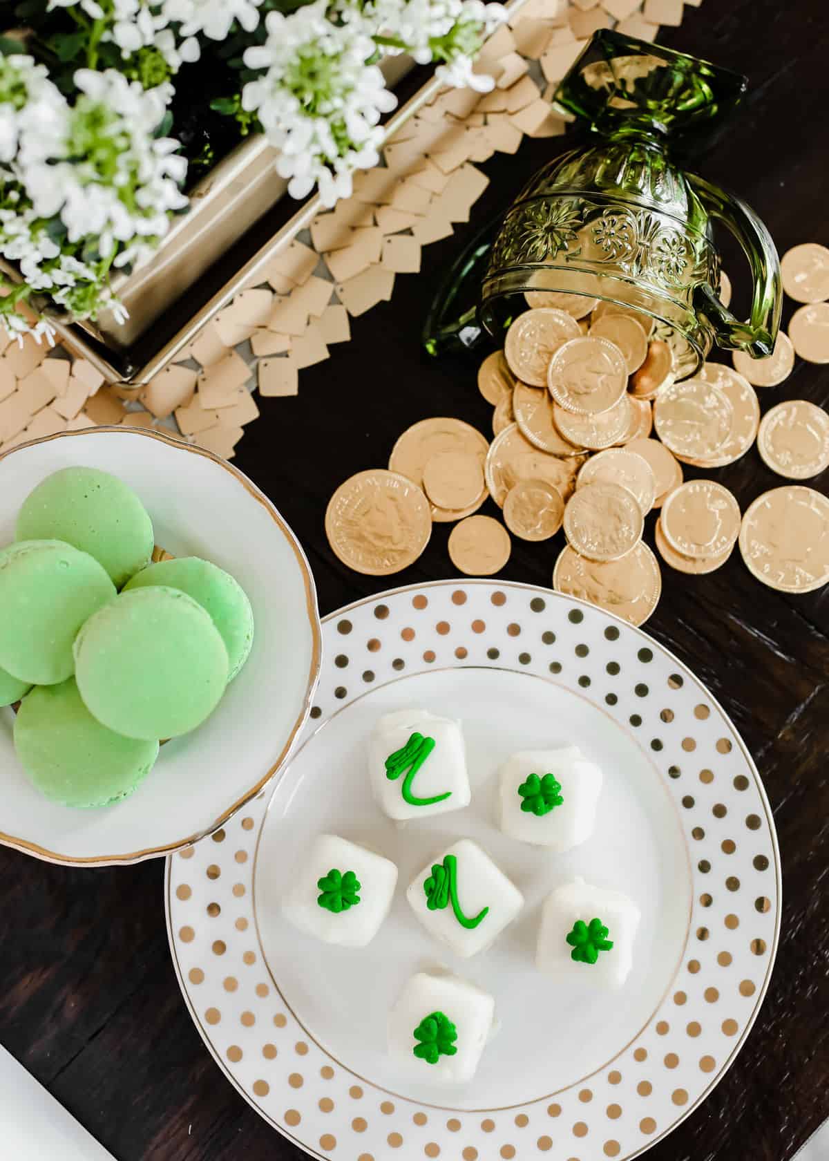 overhead view of green macarons and shamrock themed petit fours on white plates with gold coins on table.
