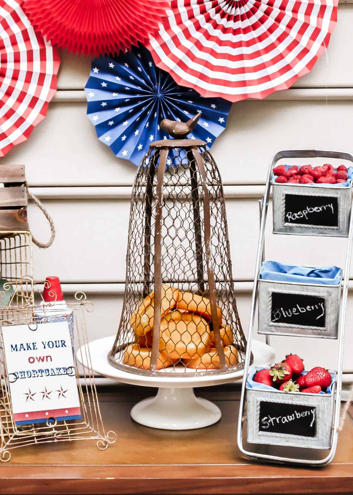 red, white, and blue decorations on table with little sponge cakes, fresh berries, and dessert table sign.