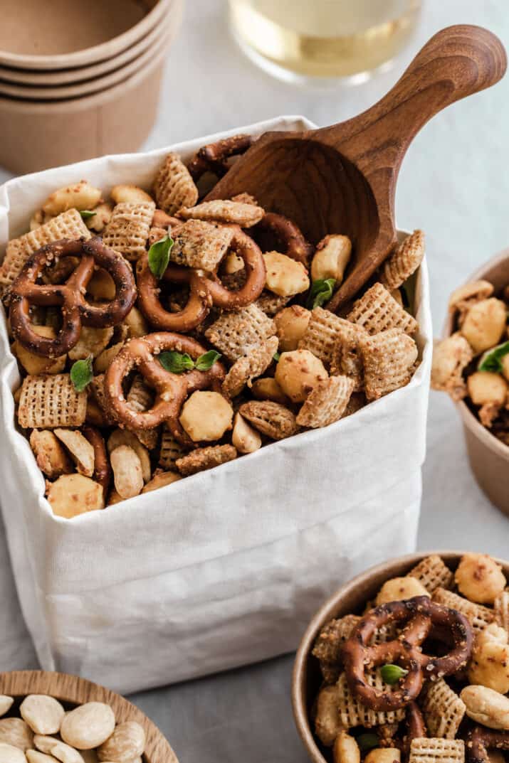 white paper bag rolled down and filled with Chex party mix with small wood scoop inside, surrounded by snack cups and drinks.