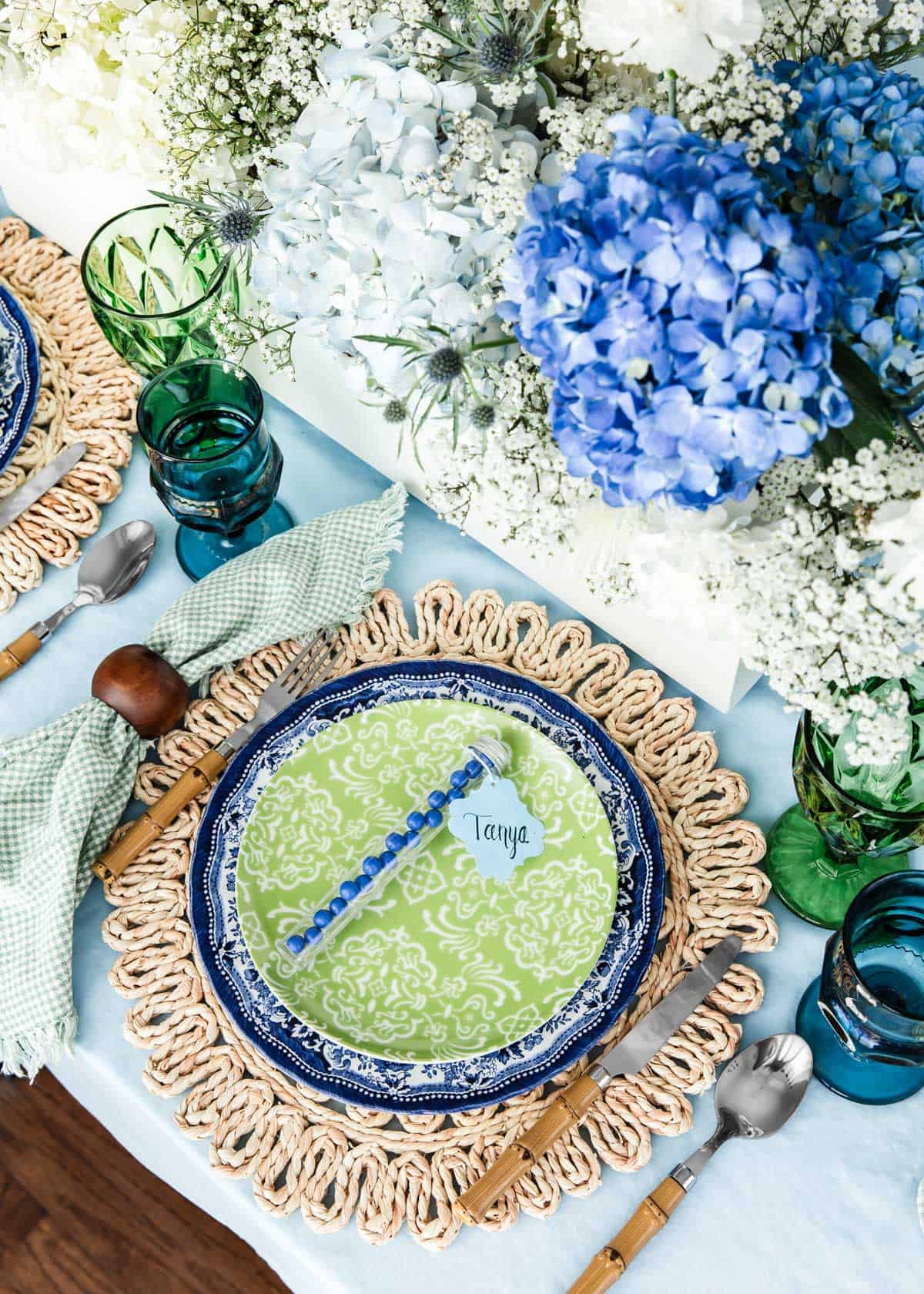 dinner table with woven place mat, blue dinner plate, green salad plate and blue and green glasses, with hydrangeas centerpiece.
