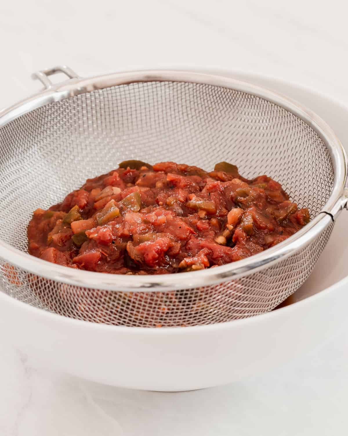 hand strainer with salsa in it, sitting over white bowl.