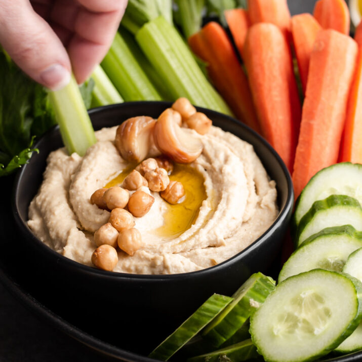 hand dipping celery into hummus in black bowl.