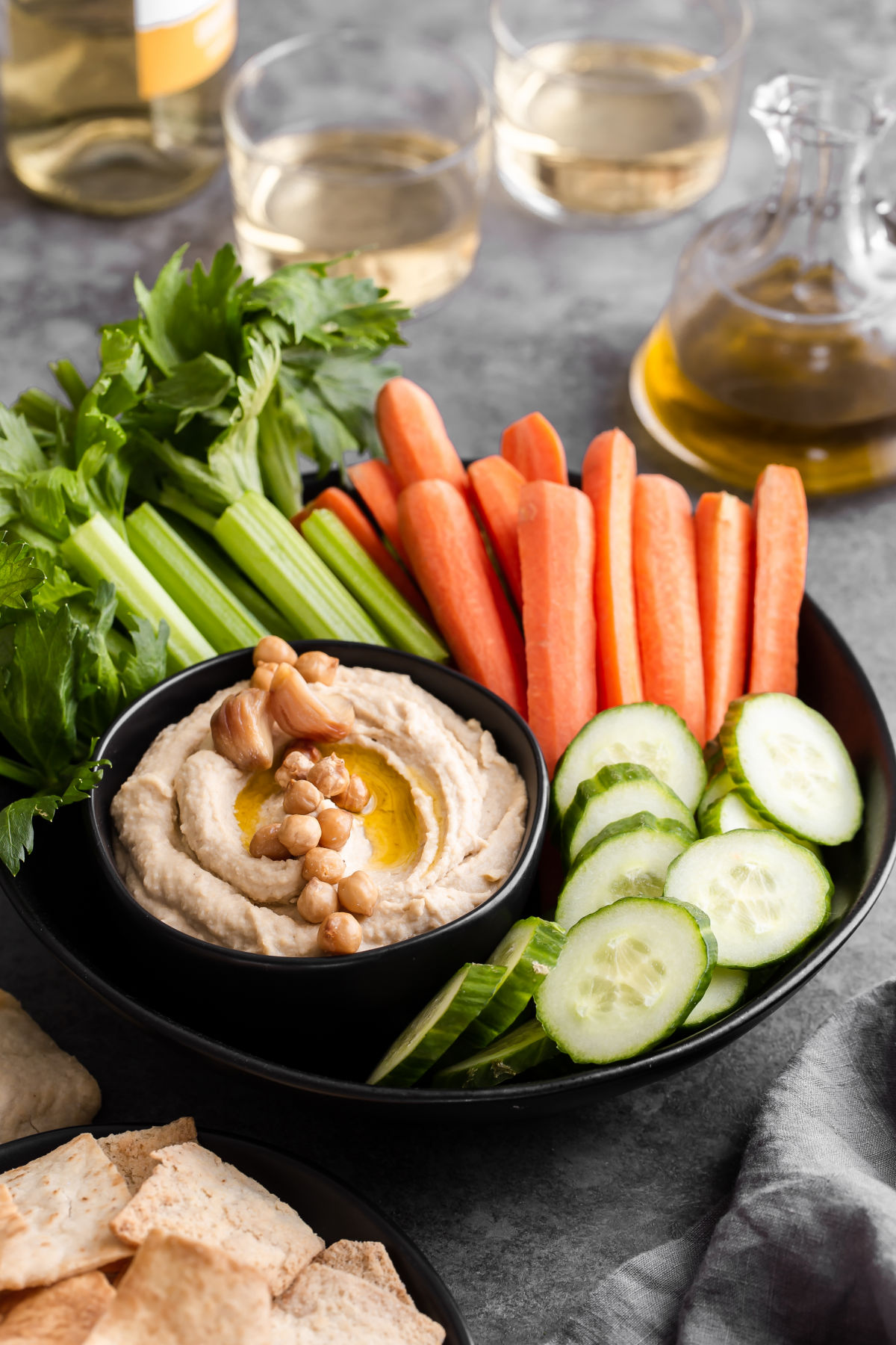 hummus in black bowl with celery, carrots and cucumber slices surrounded by wine and pita chips.