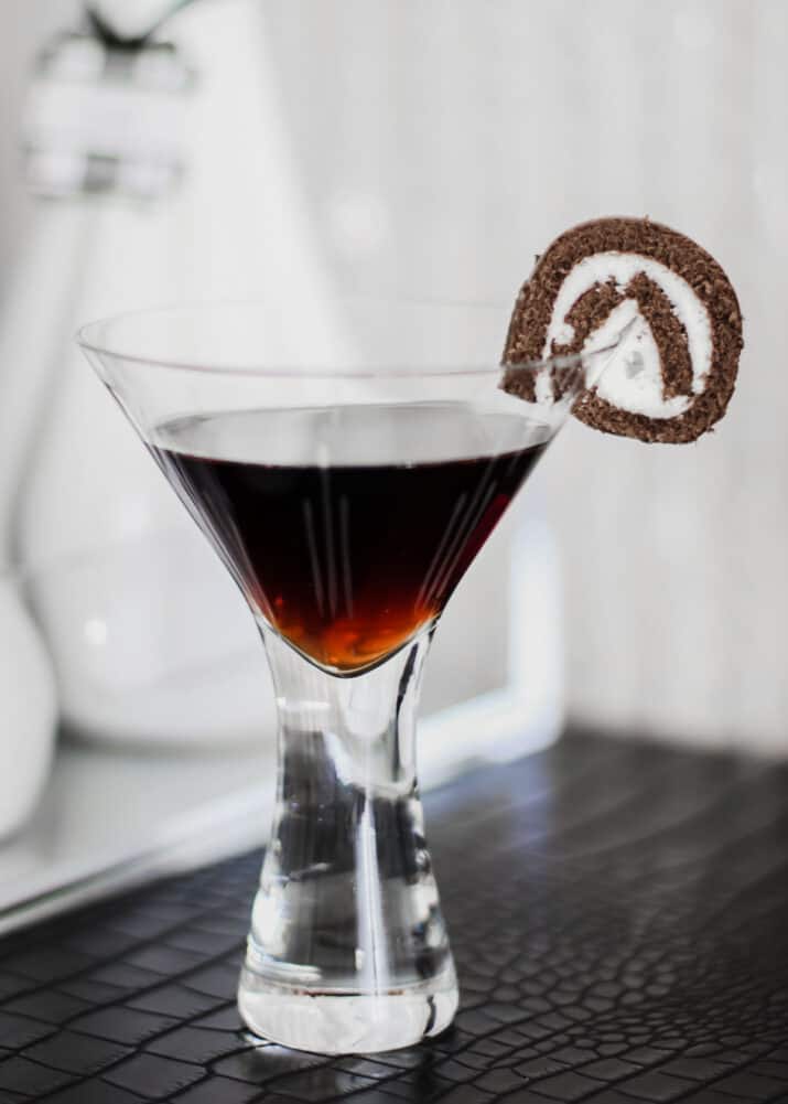 dark colored drink in martini glass with slice of rolled chocolate cake on the rim.
