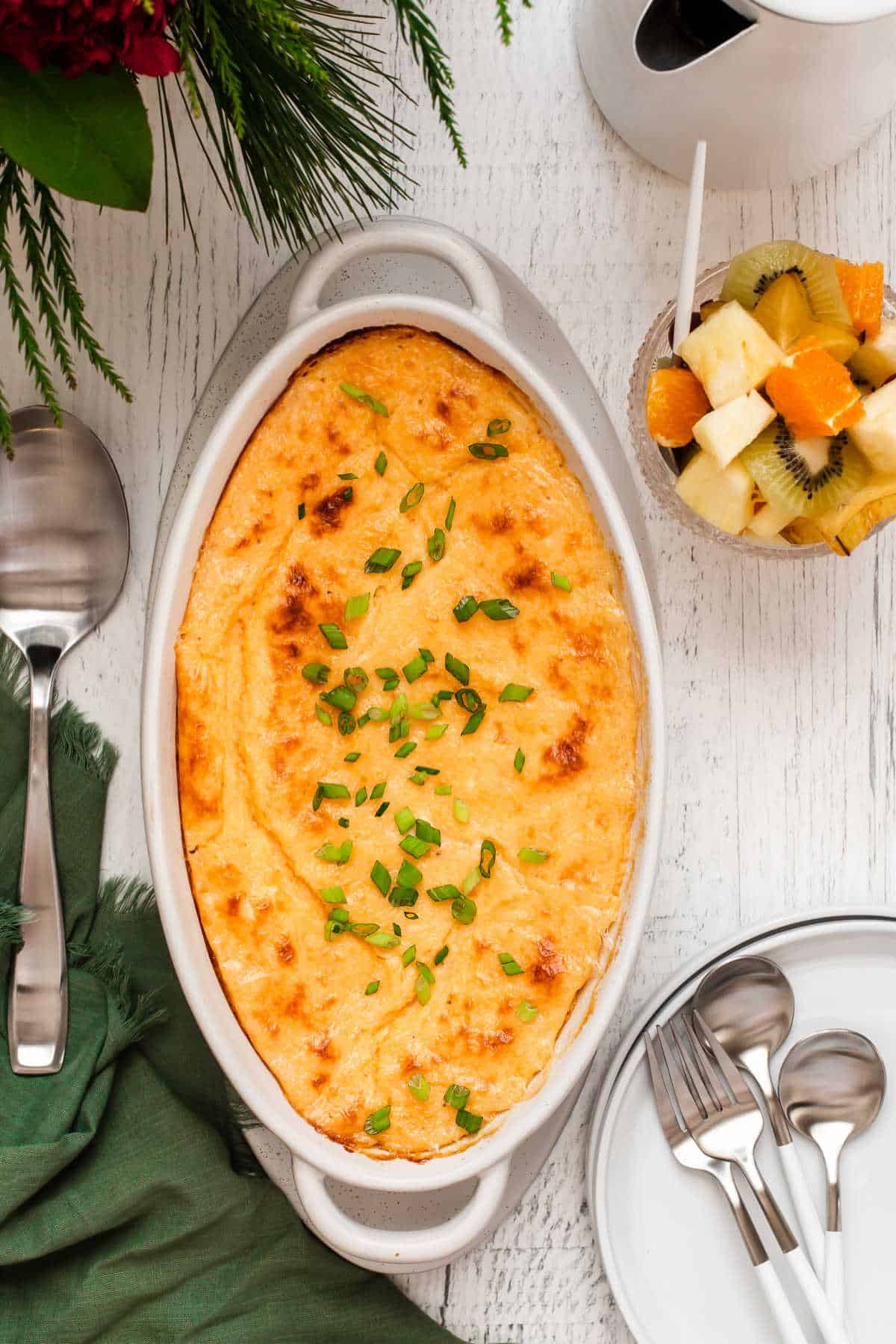 baked cheesy grits casserole on table with fruit and plates, overhead view.