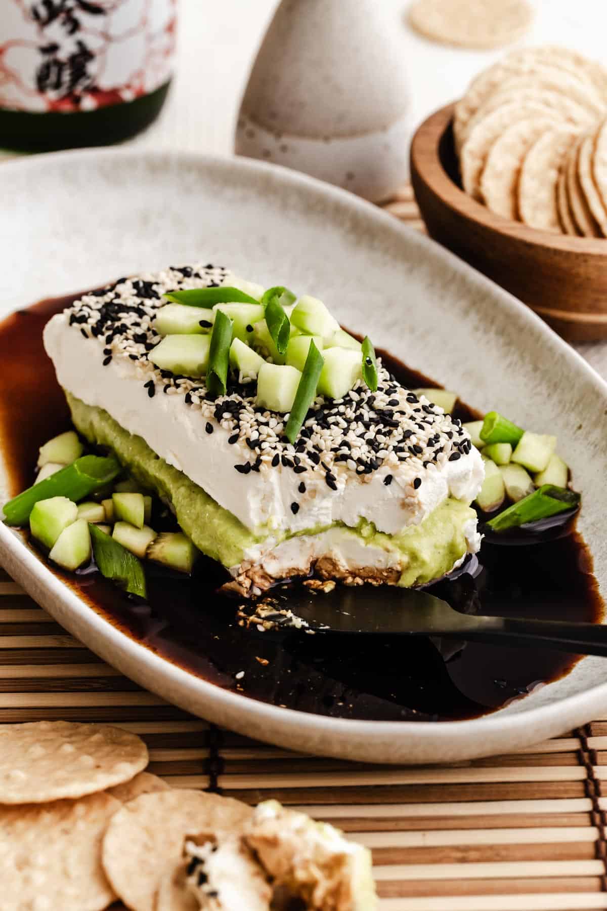 block of cream cheese with wasabi cream layered in the middle, on a plate surrounded by soy sauce and topped with sesame seeds and cucumber garnish.
