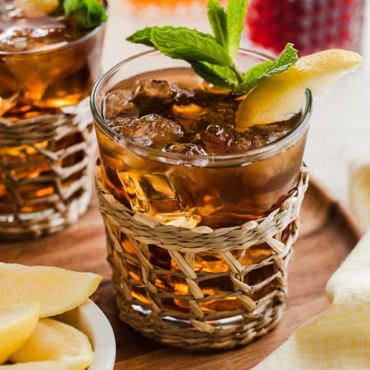 glass of bourbon iced tea with lemon and mint leaf garnish, on wood tray with bowl of lemon wedges.