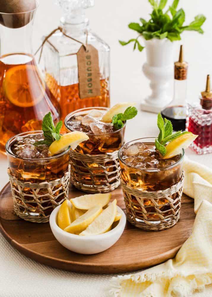 round wood tray with 3 glasses of bourbon iced tea garnished with lemon and mint, with tea pitcher and bourbon decanter in the background.