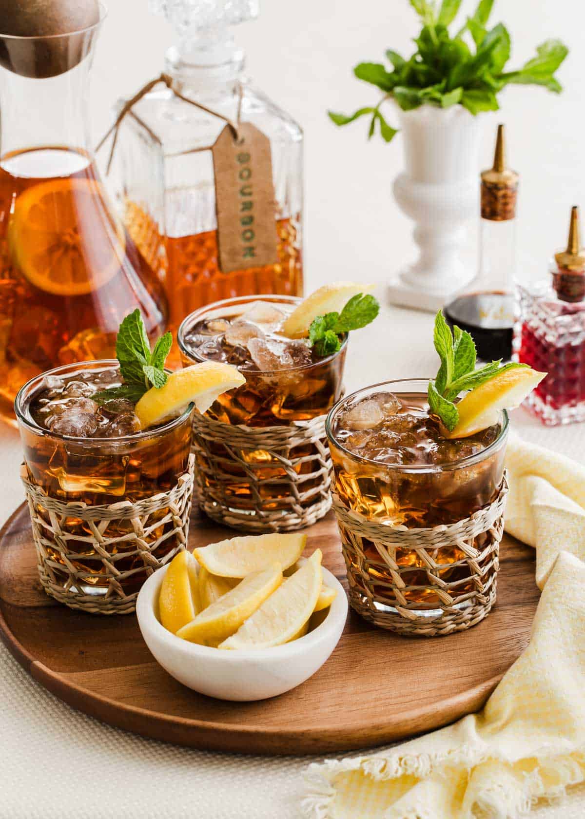 round wood tray with 3 glasses of bourbon iced tea garnished with lemon and mint, with tea pitcher and bourbon decanter in the background.
