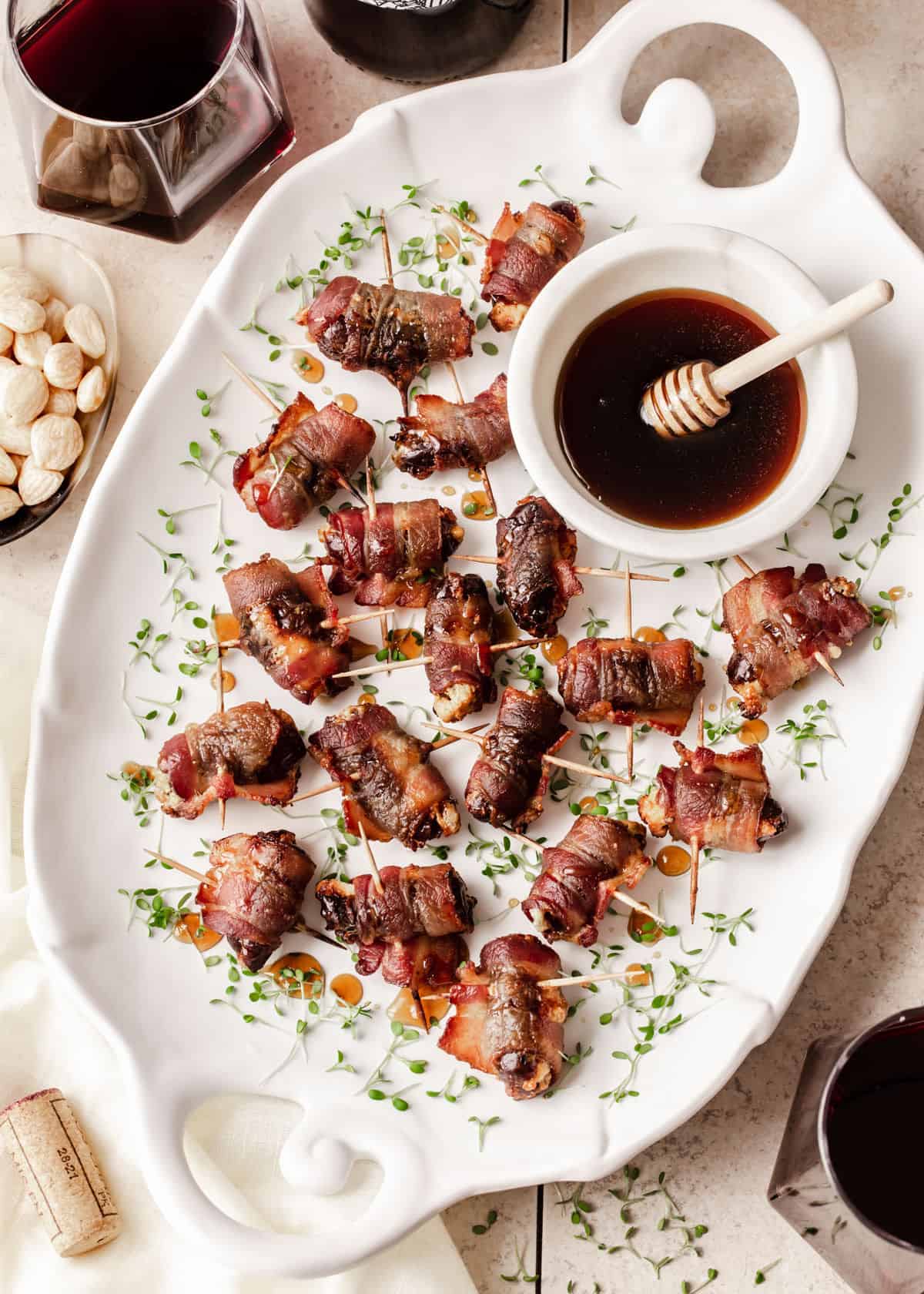 white platter filled with bacon wrapped dates appetizers with small bowl of honey, surrounded by nuts and wine glasses, overhead view.