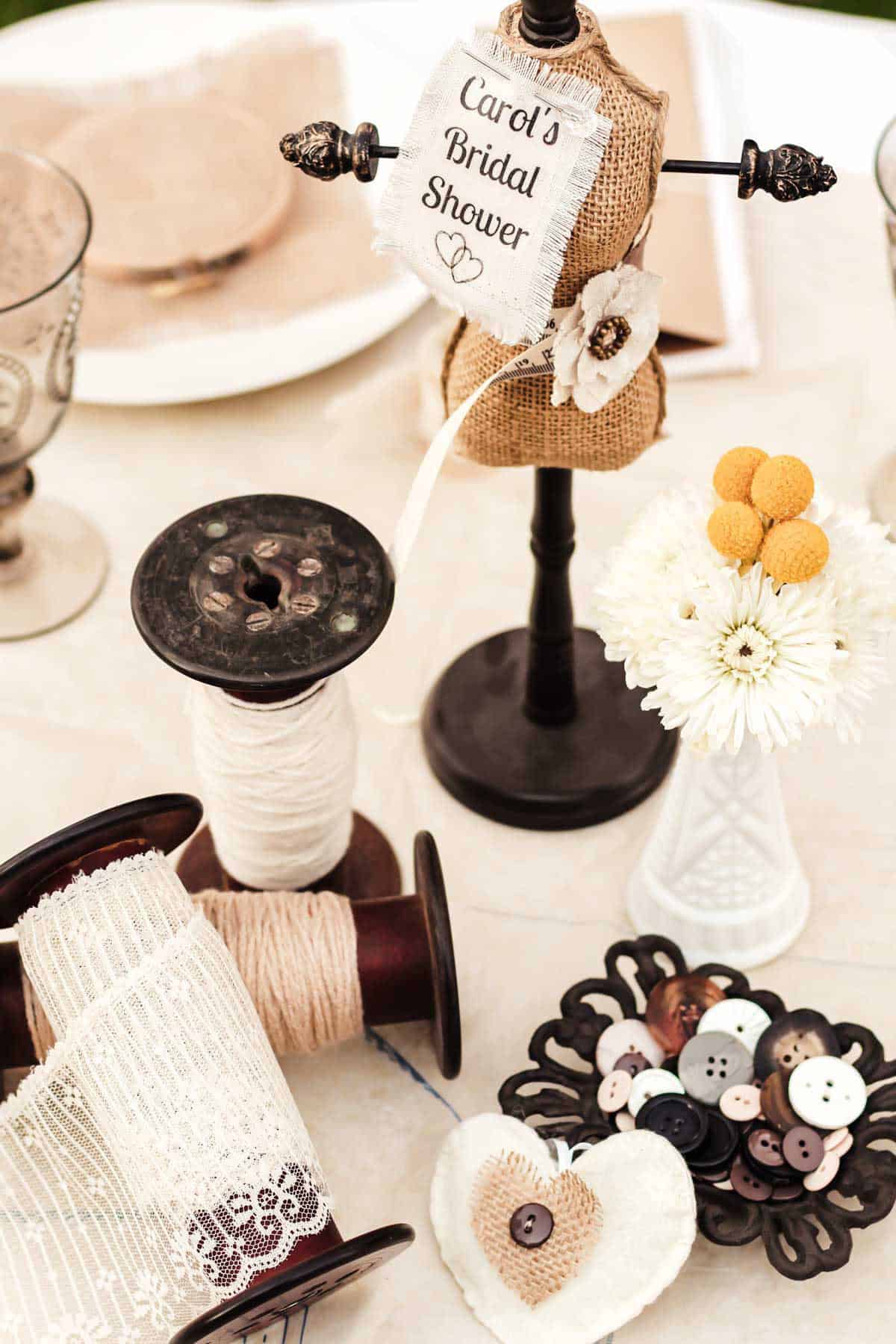 shabby chic table centerpiece with large vintage spools, dish of buttons, and mini dress form with table sign attached.