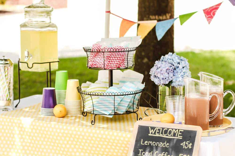 outdoor table set up for a lemonade stand with colorful fabric, drink dispenser, and snacks.