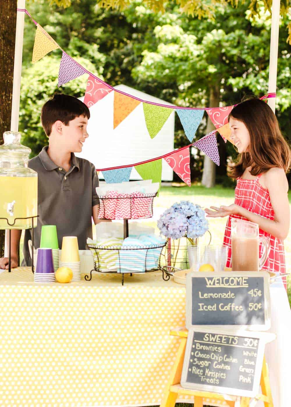 boy and girl standing behind a colorful lemonade stand.