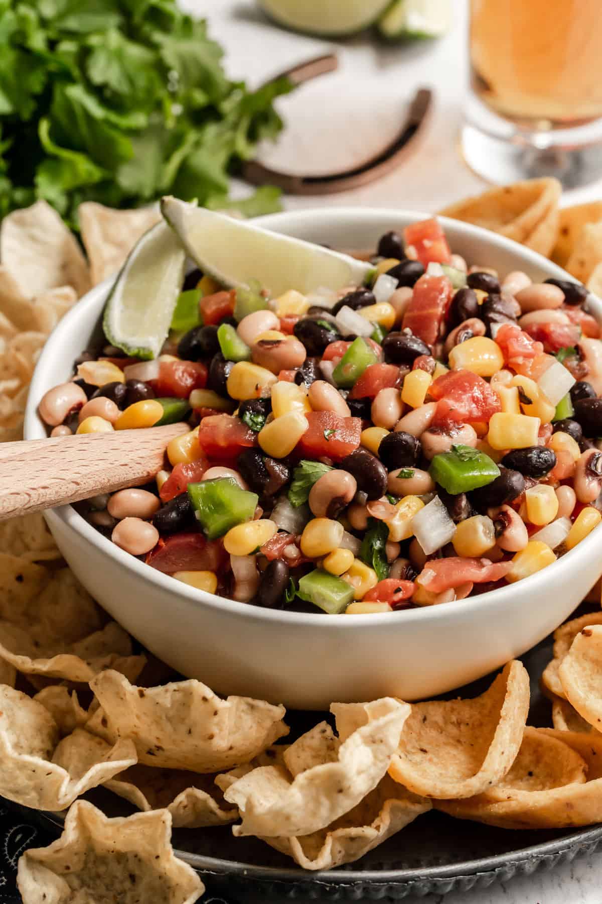 cowboy salad with black beans, pinto beans, corn and tomatoes in white bowl with wood spoon inside and garnished with lime wedges.