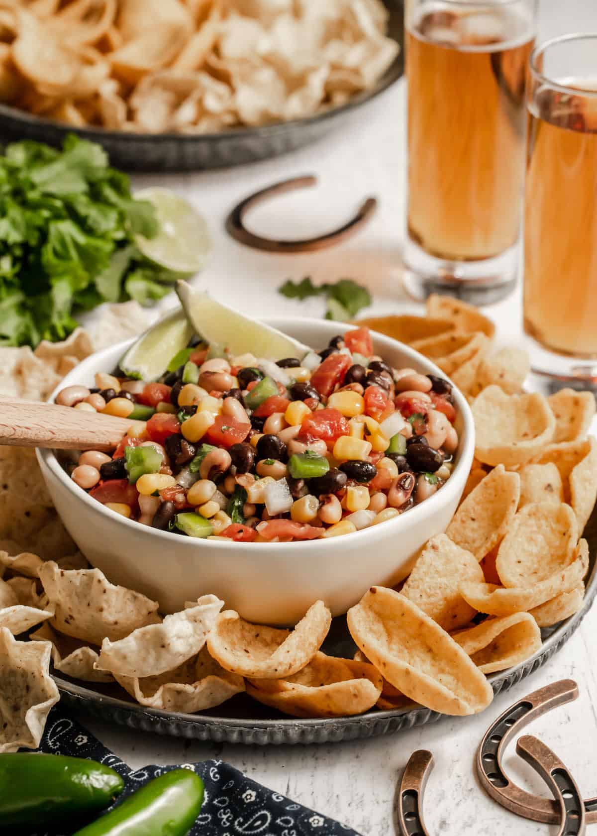 redneck caviar in white bowl surrounded by tortilla and Fritos scoops, with beer glasses and small horseshoe decorations on table.