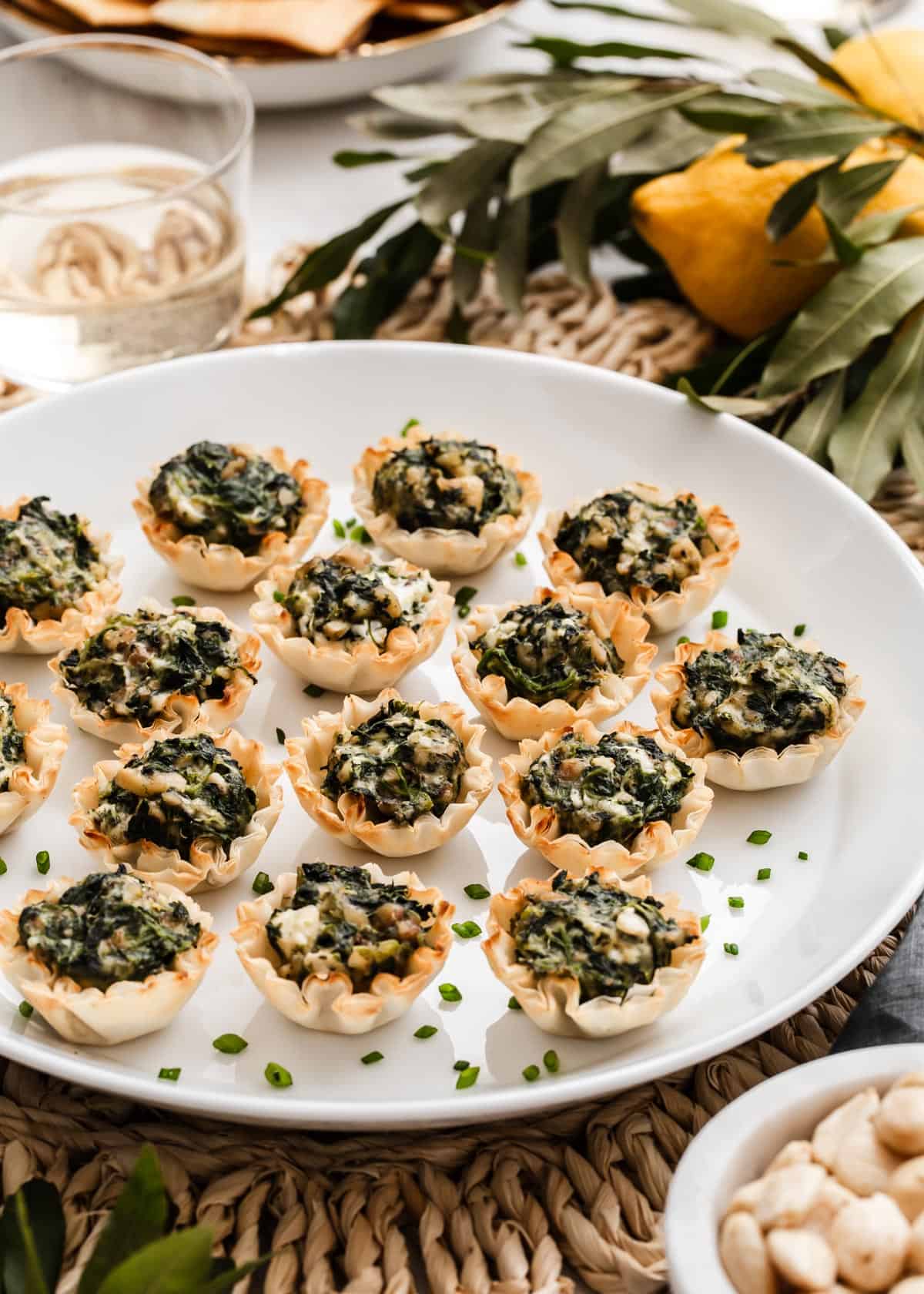 phyllo shells appetizers with spanakopita filling, on white platter with wine and lemons in background.
