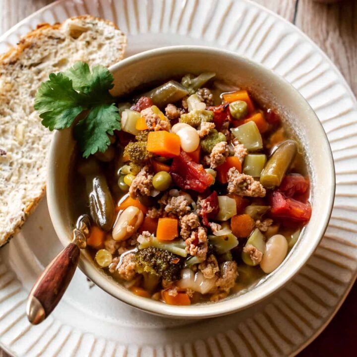 ground turkey vegetable soup in cream bowl on cream plate with bread slice on the side, overhead view