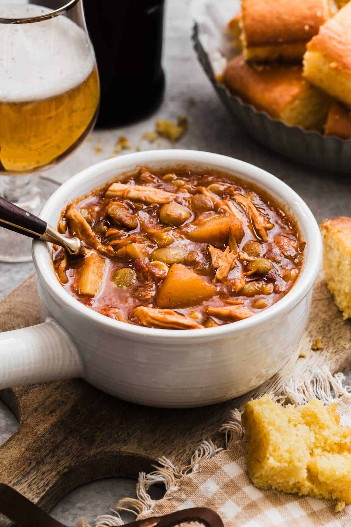 Brunswick stew in white bowl with handle, spoon inside, cornbread and beer glass in background.