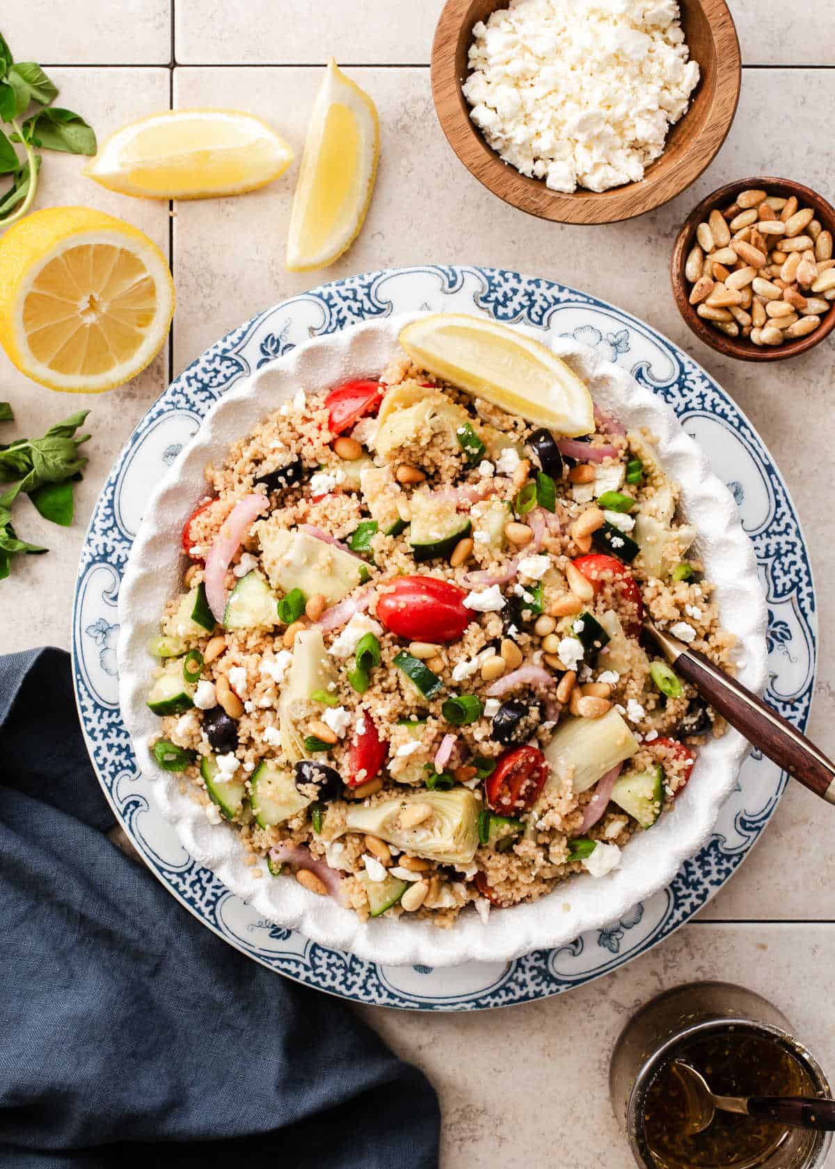 round bowl filled with couscous salad full of veggies, layered on blue and white plate surrounded by lemons, dressing and pine nuts.