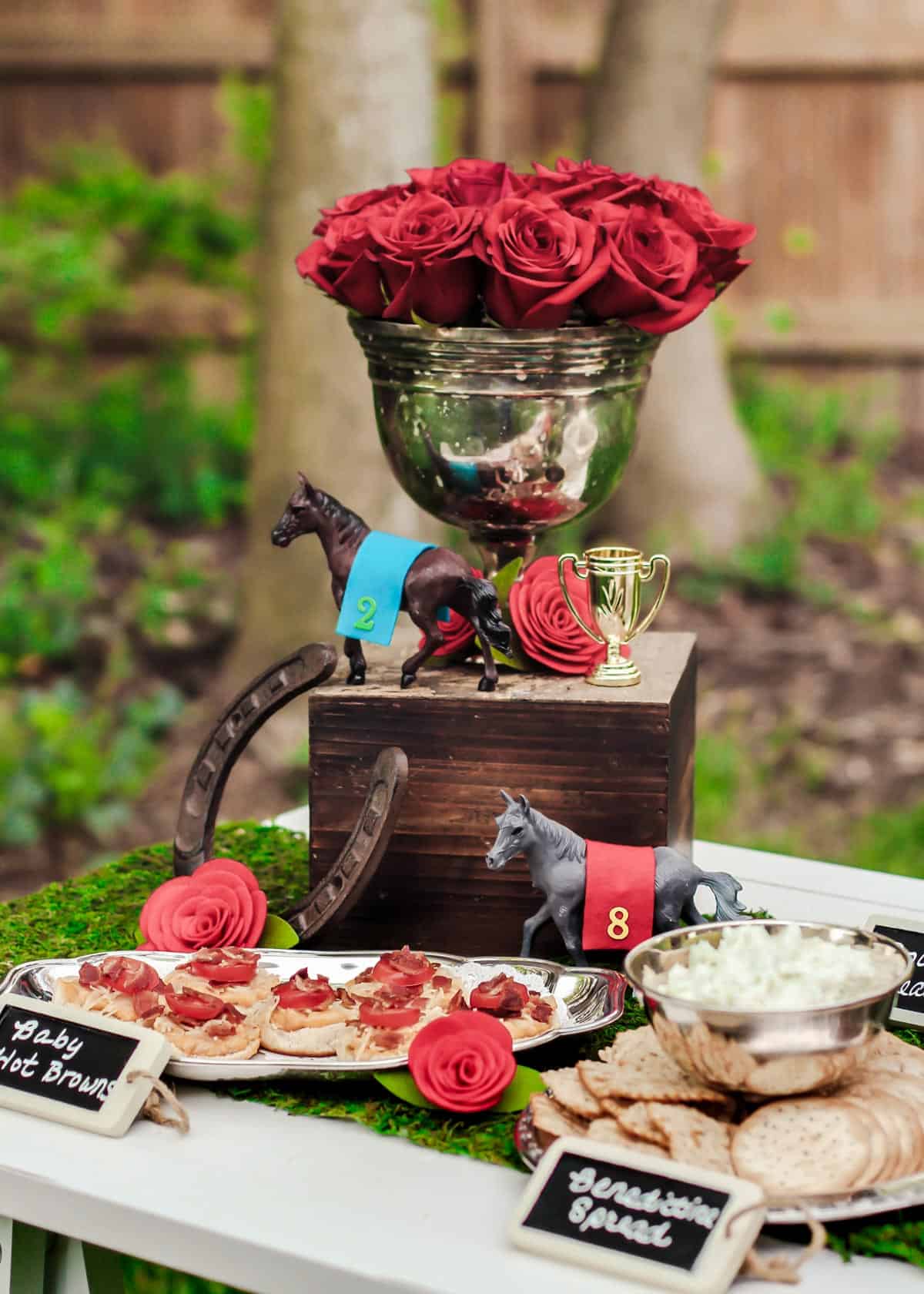 Kentucky derby party set up outdoors with roses in silver urn, horse decor, and food on table.