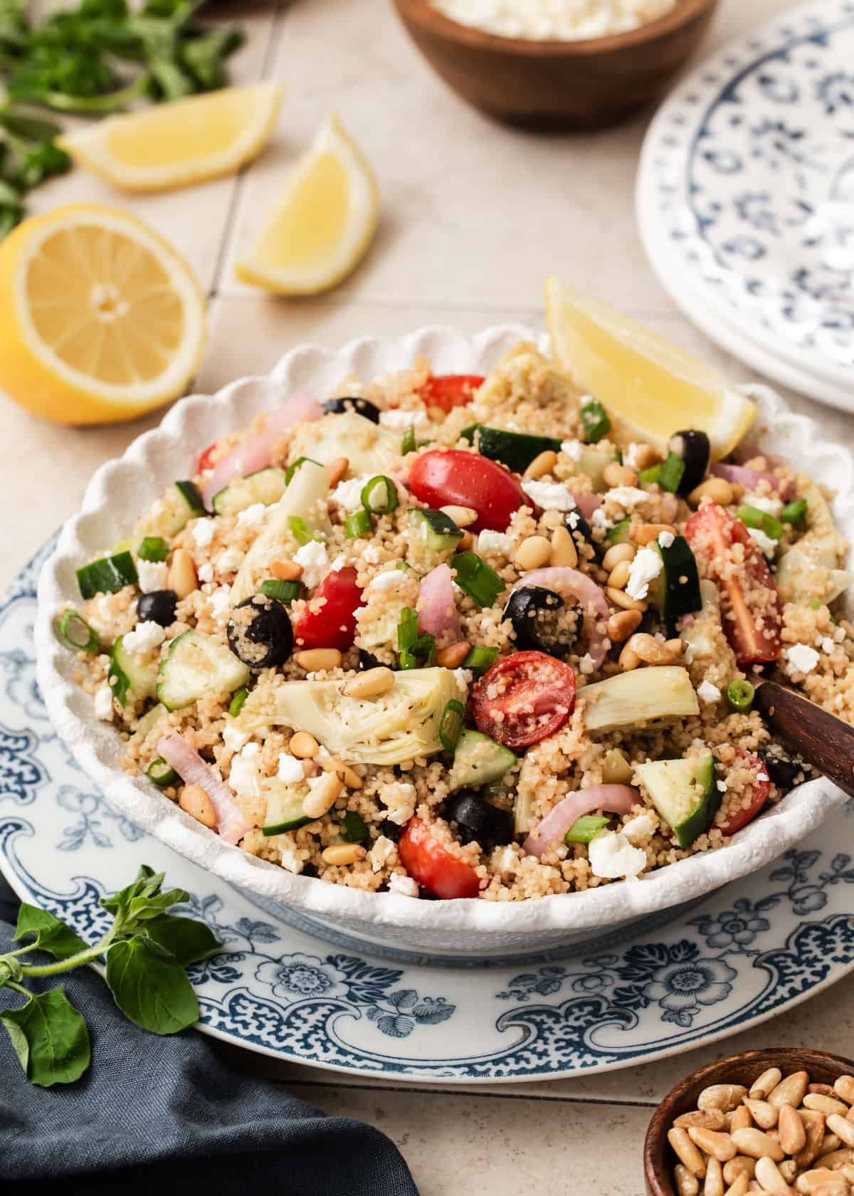 round bowl filled with couscous salad full of veggies, layered on blue and white plate surrounded by lemons, pine nuts and green herbs.