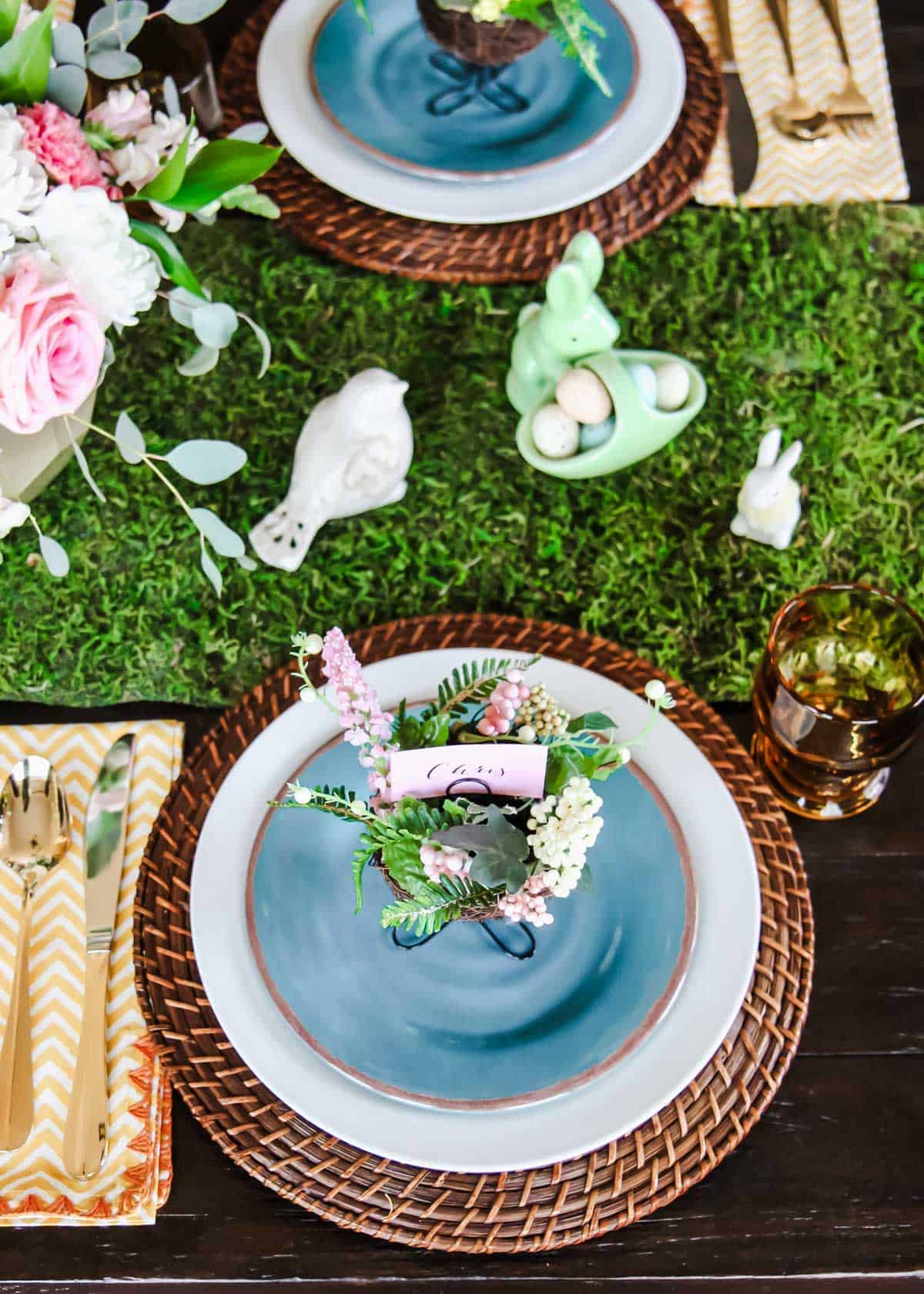 Easter tablescape with moss runner, white and blue dishes, rattan place mat, and bunny figurines.