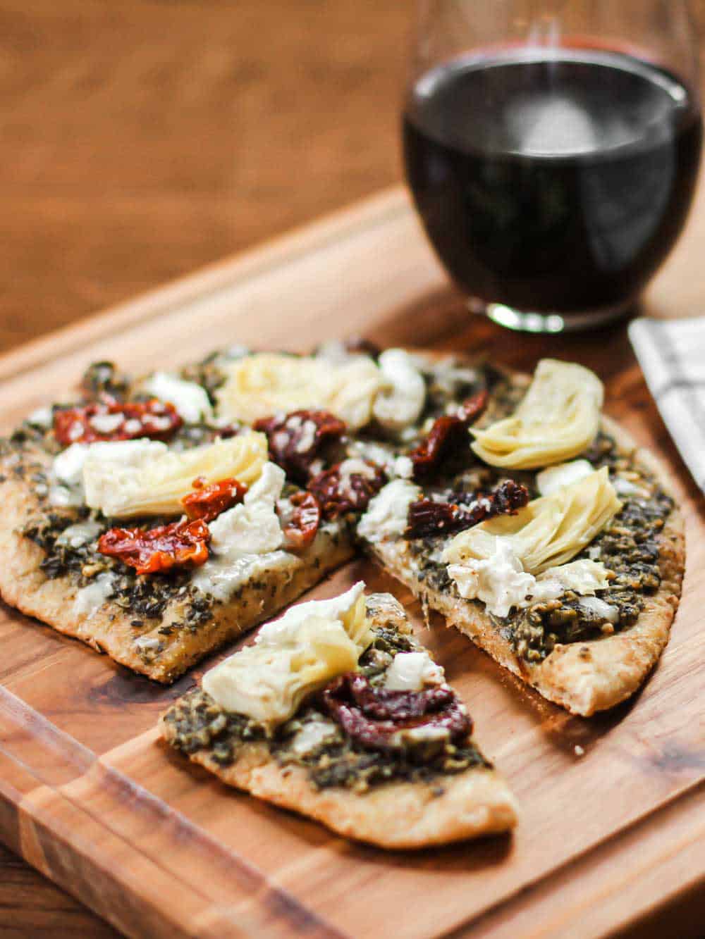 pizza sliced on wood board, with artichokes, pesto, and sundried tomatoes.