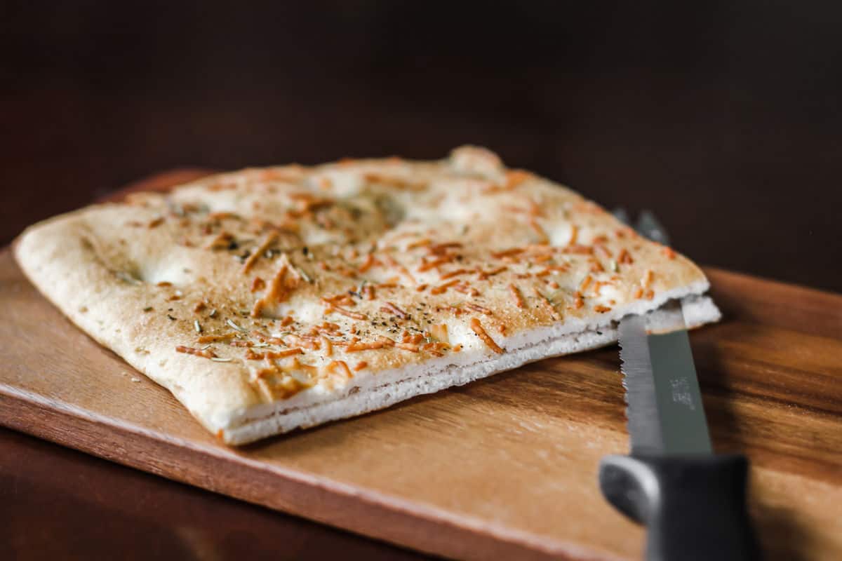 focaccia bread on wood board with knife cutting through the middle.