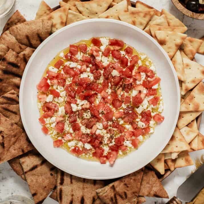round dish with diced tomatoes, feta cheese and olive oil dip inside, surrounded by pita chips.