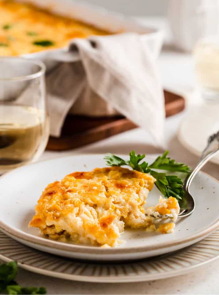 cheesy corn recipe scooped onto white plate with casserole dish in background, side view.