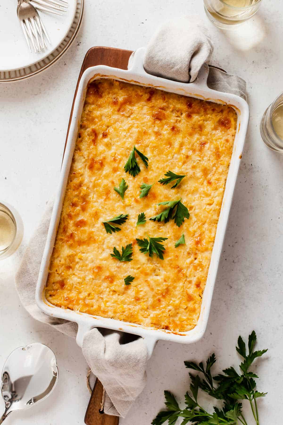 yellow corn casserole in a white baking dish on white table with plates and drink glasses, overhead view.