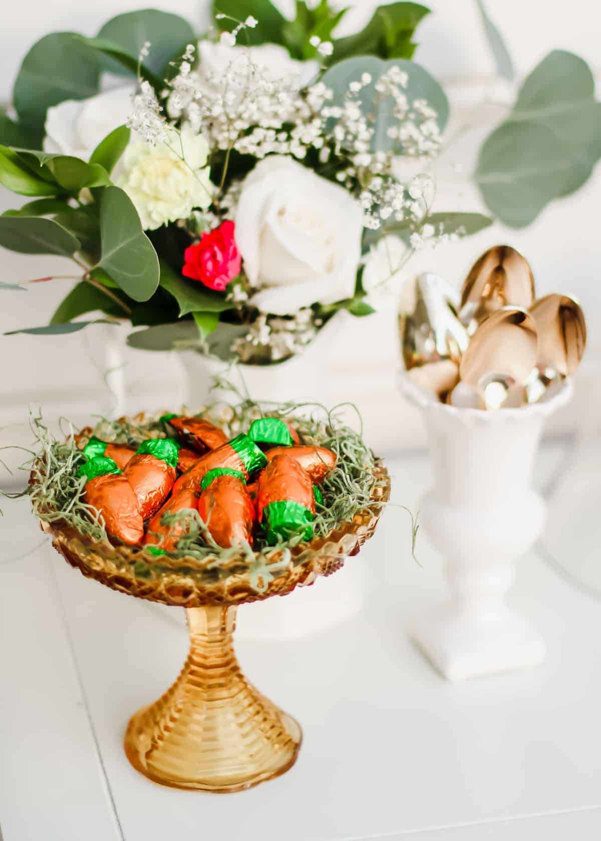 flower arrangement with dish filled with Easter candy, and white jar holding gold spoons.