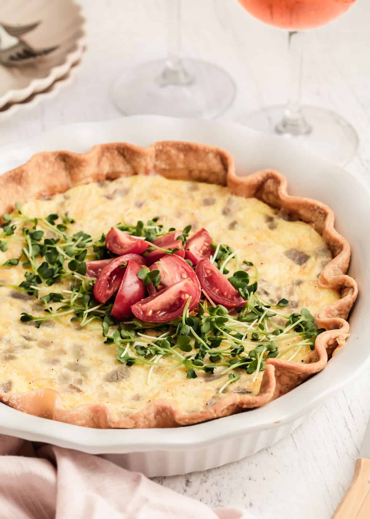 quiche topped with microgreens and cut tomatoes, in white dish sitting on white table with stem glass in the background.