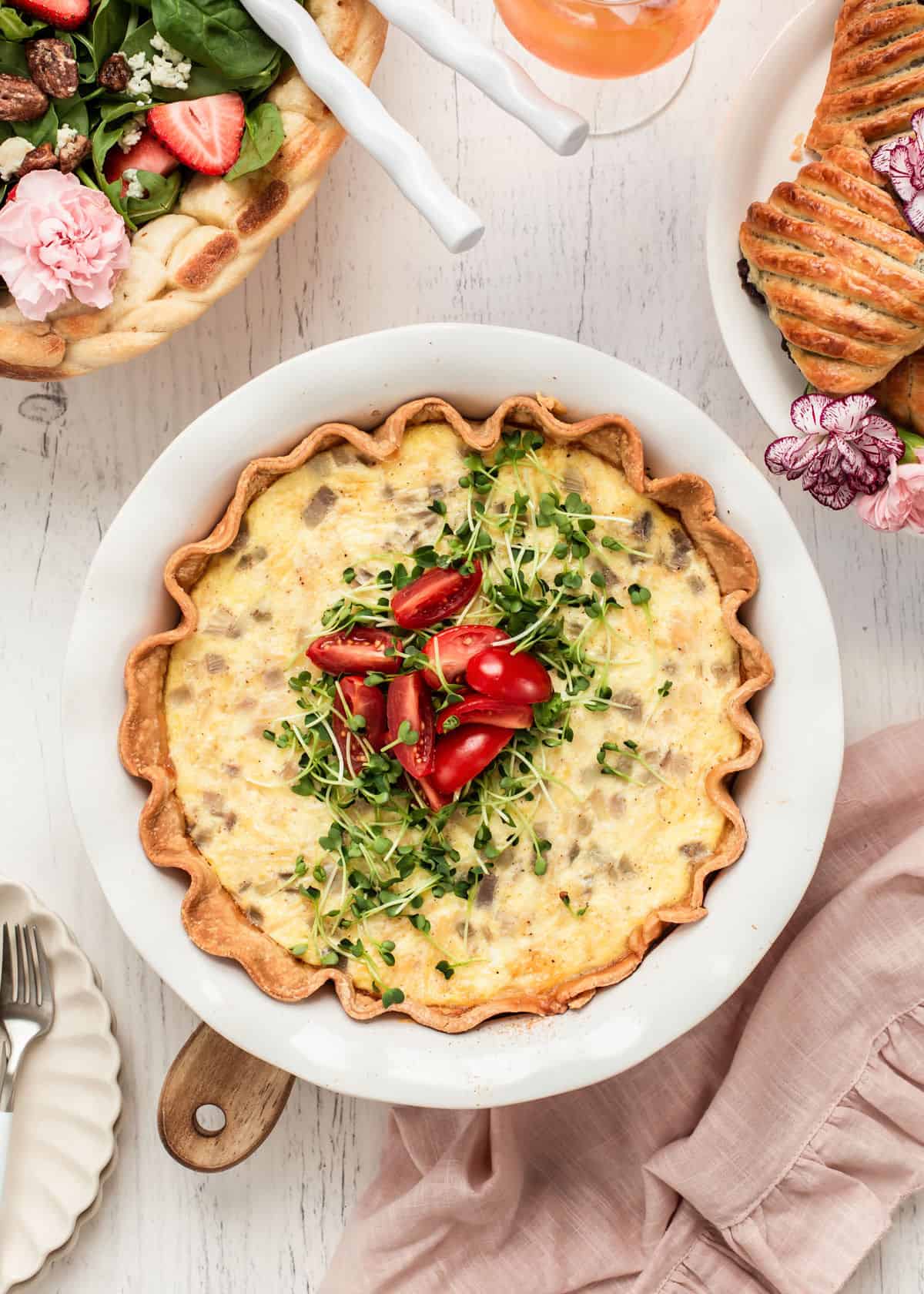 quiche garnished with microgreens and cherry tomatoes, sitting on white table surrounded by other brunch foods.