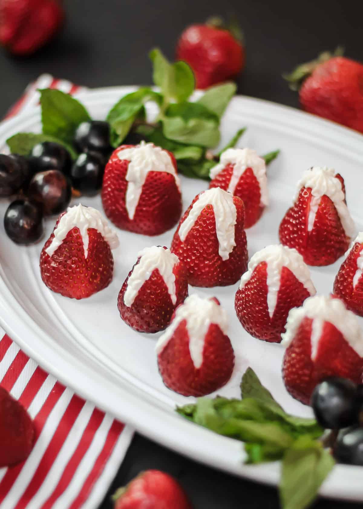 strawberries stuffed with cheesecake filling, on white platter garnished with grapes and mint leaves.