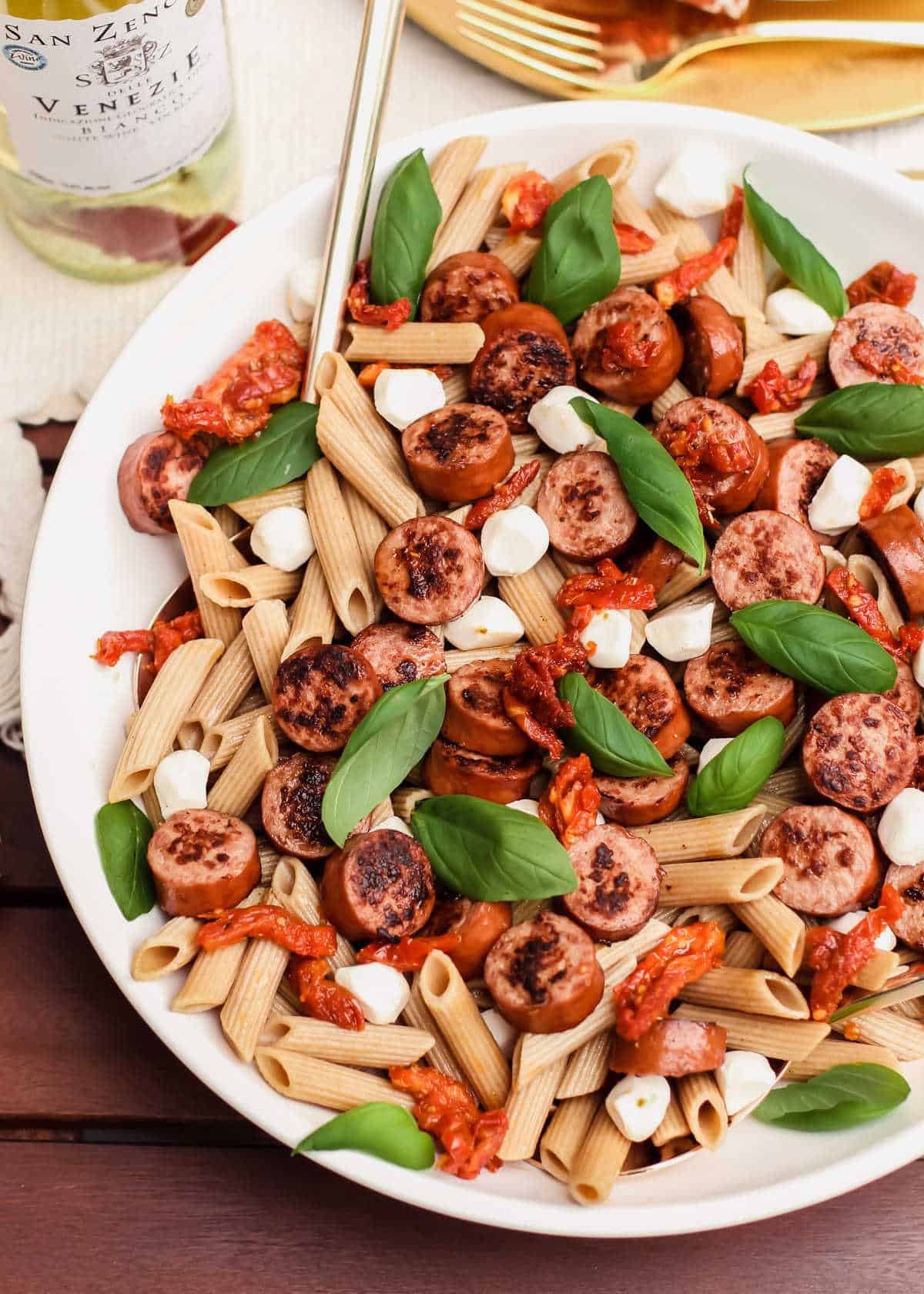 round bowl of penne pasta with sliced sausage, sundried tomatoes, mozzarella balls, and basil leaves.