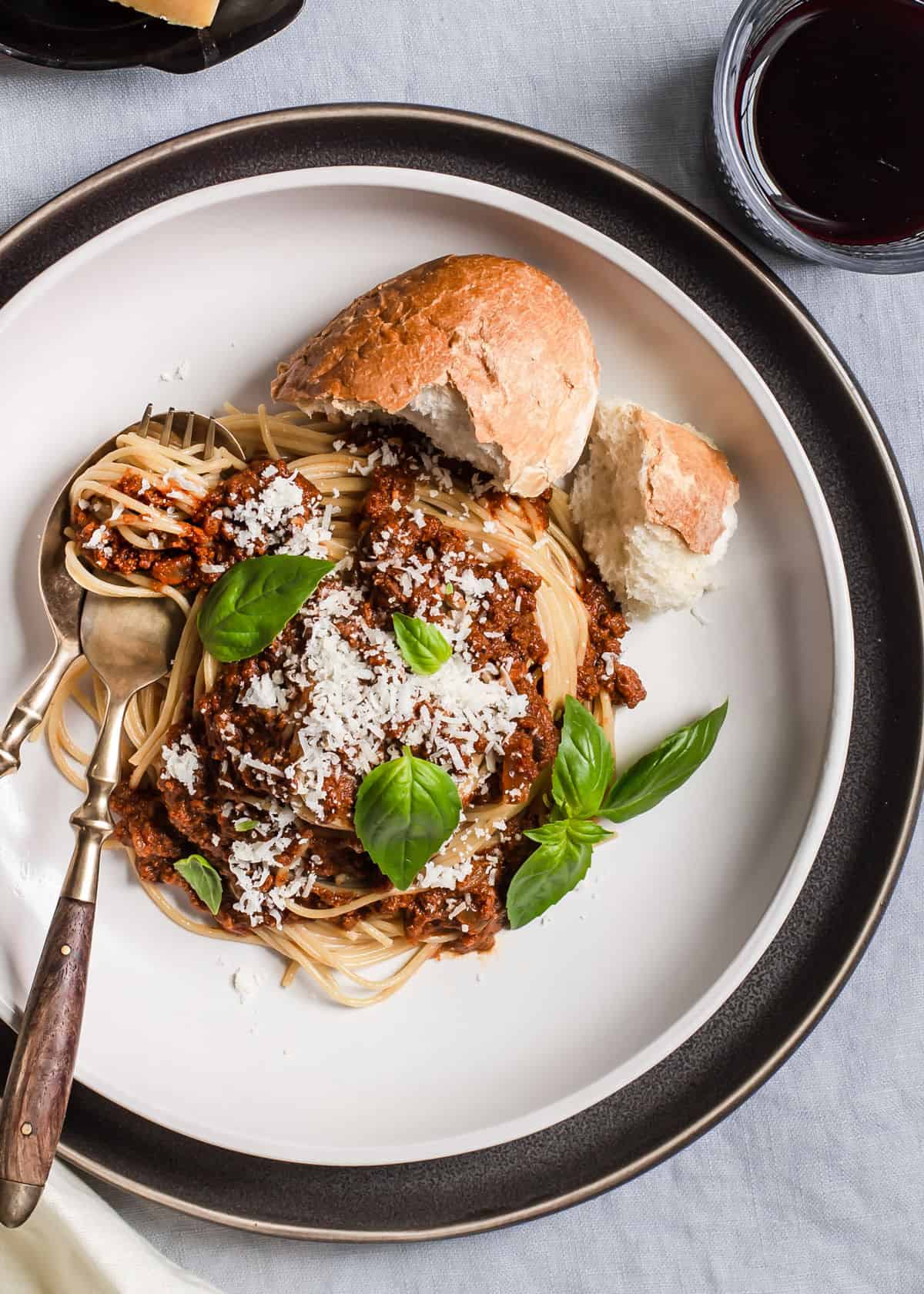 overhead view of spaghetti and meat sauce on white plate, garnished with basil leaves and bread on the side.