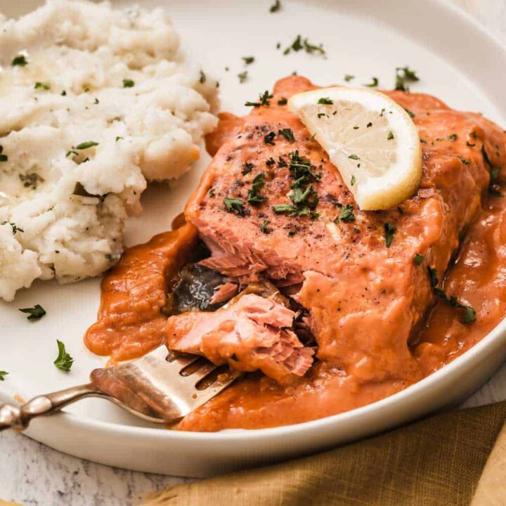 salmon fillet in red sauce on plate with fork taking a bite, and mashed potatoes on the side.