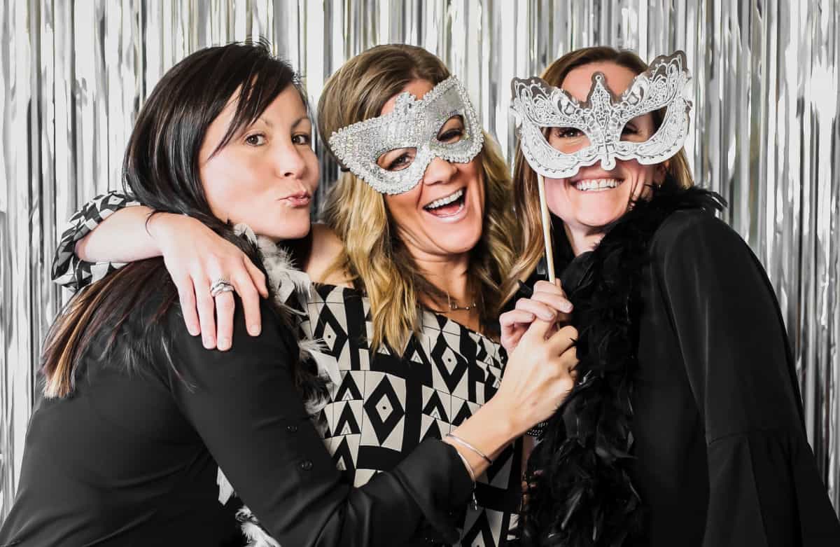 three ladies posing with masquerade masks and silver fringe backdrop.