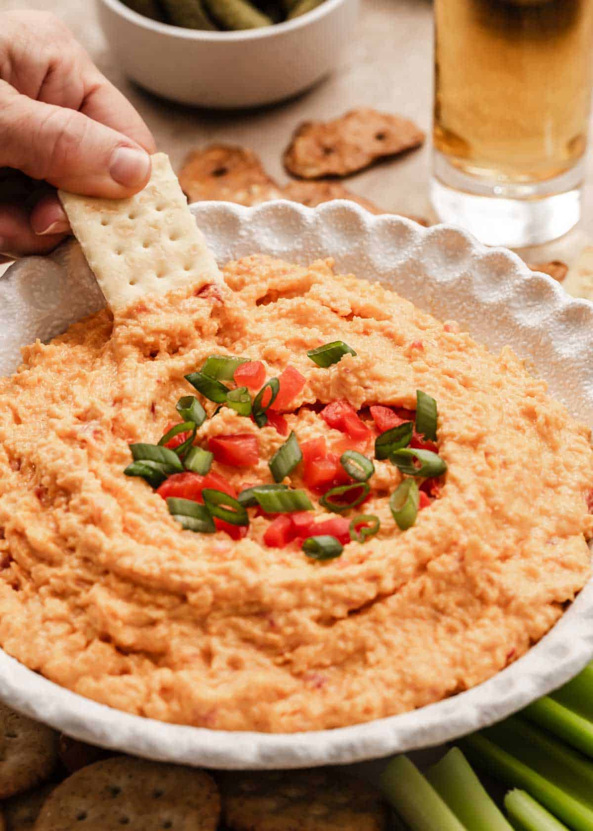 pimento cheese in white bowl with hand dipping a cracker into.