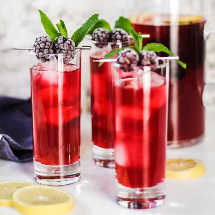 three tall glasses filled with red drink on ice garnished with blackberries and mint, with pitcher in background.